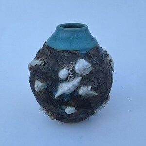 May include: A handcrafted ceramic vase with a textured, dark brown body and a turquoise blue rim. The vase is adorned with white seashell and barnacle embellishments, creating a coastal or nautical theme. The vase has a round shape.