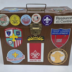 May include: A brown metal storage box with a wood grain finish, featuring a gold-colored metal latch and handle. The exterior is covered in various colorful stickers related to scouting, including emblems, text labels, and a portrait of Baden-Powell.
