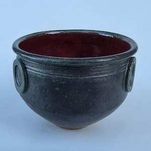 May include: A handmade ceramic bowl with a dark exterior and a deep red interior. The bowl features two decorative circular handles and a green rim. The bowl's exterior has a textured, matte finish.