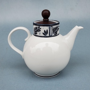 May include: White ceramic teapot with a round body, a curved handle, and a spout. The lid is dark brown with a matching finial. The rim of the lid features a navy blue band with a leaf design. A navy blue band encircles the base.