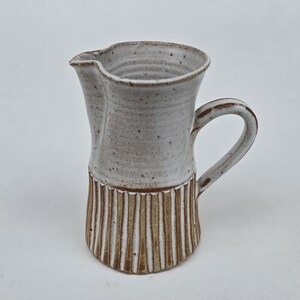 May include: A handmade ceramic pitcher with a cream-colored upper section and a textured, vertical-striped brown base. The pitcher has a curved handle and a spout for easy pouring. The stoneware piece is ideal for serving beverages.