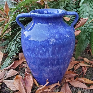 Op de afbeelding: Een blauwe keramische vaas met twee handvatten. De vaas heeft een gevlekt blauw glazuur met witte strepen. Hij staat op een bed van bruine bladeren.