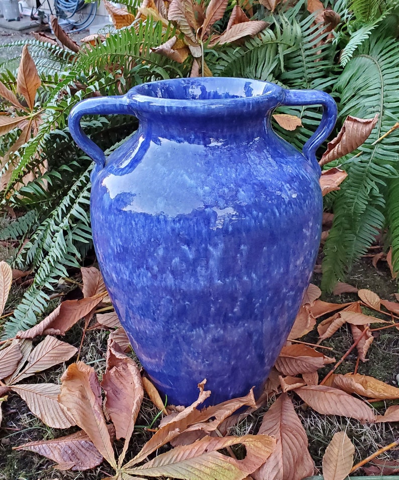 Op de afbeelding: Een blauwe keramische vaas met twee handvatten. De vaas heeft een gevlekt blauw glazuur met witte strepen. Hij staat op een bed van bruine bladeren.