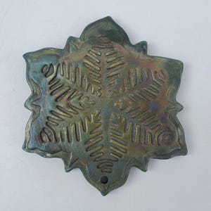 May include: A decorative, snowflake-shaped ornament with a metallic finish. The ornament features a detailed snowflake design with a hole at the bottom for hanging. The color is a mix of green, blue, and bronze.