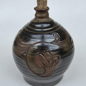 May include: A round, dark brown and black glazed ceramic jug with a narrow neck and a cork stopper. The surface features an incised, stylized floral pattern that reveals the lighter brown clay underneath the dark, glossy glaze finish.