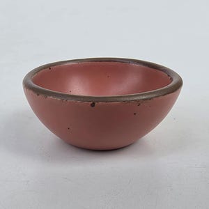 May include: A small, round ceramic bowl with a matte dusty pink interior and exterior. The bowl features a thick, dark brown rim that contrasts with the pink body. The surface of the bowl has a slightly textured finish with small, visible dark speckles.
