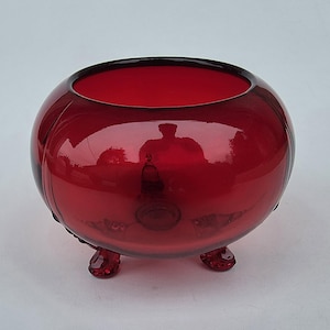 May include: A ruby red glass bowl with a rounded shape and three small feet. The bowl has a smooth, reflective surface and a wide opening. The glass appears to be a deep, rich red color, and the bowl is likely a decorative item.