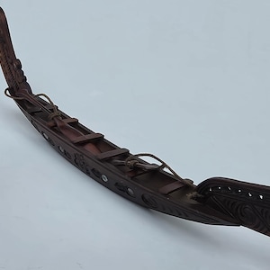 May include: A dark brown wooden model canoe featuring intricate carved patterns along the hull and bow. The vessel includes inlaid iridescent shell accents and two thin leather cords tied across the open deck area, showcasing detailed craftsmanship.