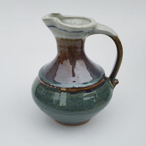 May include: A small, hand-thrown ceramic pitcher with a wide, rounded body and a short, curved spout. The pitcher is glazed in shades of green, brown, and white.