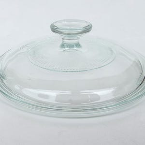 May include: A clear glass lid with a knob handle. The lid is round with a slightly raised center section featuring a radial design. The edges are smooth and curved, suitable for cookware.