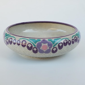 May include: A decorative ceramic bowl with a crackle glaze finish. The bowl features a purple rim and a band of teal and purple floral designs around the exterior. A central purple flower with a pink center is also present.