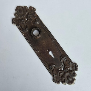 May include: A rectangular dark bronze door plate featuring ornate scrollwork at the top and bottom ends. The metal plate includes a central circular opening for a door knob and a lower keyhole opening, with visible signs of aged patina and wear.
