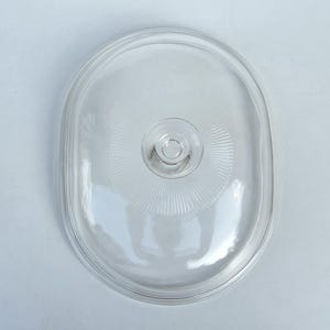 May include: A clear, oval-shaped glass lid featuring a central knob handle. The glass surface displays a subtle, radiating sunburst pattern emanating from the center, with a smooth, raised outer rim designed to fit securely onto a matching container.