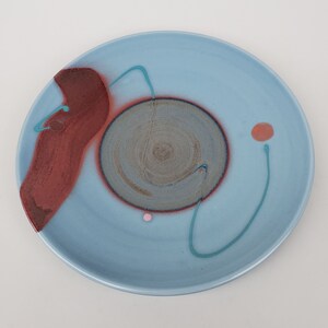 Può includere: Un piatto in ceramica blu chiaro con disegni astratti in tonalità di marrone, rosa e verde acqua. Il piatto presenta un grande disegno circolare al centro con motivi a spirale.