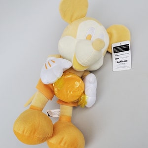 Puede incluir: Juguete de peluche amarillo de Mickey Mouse sosteniendo una moneda amarilla. El juguete lleva un atuendo amarillo y blanco. El juguete tiene una cara blanca con ojos negros y una nariz negra. El juguete tiene una etiqueta adjunta a su oreja derecha que dice "Disney SEGA Prize USA!"