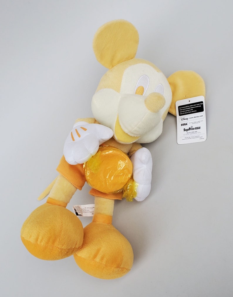 Puede incluir: Juguete de peluche amarillo de Mickey Mouse sosteniendo una moneda amarilla. El juguete lleva un atuendo amarillo y blanco. El juguete tiene una cara blanca con ojos negros y una nariz negra. El juguete tiene una etiqueta adjunta a su oreja derecha que dice "Disney SEGA Prize USA!"