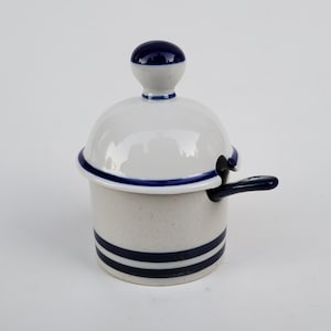 May include: A white ceramic sugar bowl with a lid and spoon. The bowl has two navy blue stripes around the base and a navy blue rim. The lid has a navy blue knob and a navy blue rim. The spoon is navy blue.