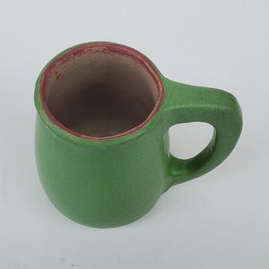 May include: A small, green ceramic mug with a rounded body and a handle. The interior is a light brown color, and the rim is trimmed with a reddish-brown hue. The mug is designed for beverages.