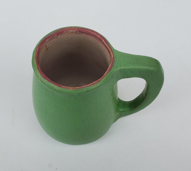 May include: A small, green ceramic mug with a rounded body and a handle. The interior is a light brown color, and the rim is trimmed with a reddish-brown hue. The mug is designed for beverages.