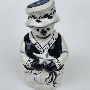 May include: A ceramic snowman figurine with a white and navy blue color scheme. The snowman wears a hat and vest, and holds a star. The figurine has a crackle glaze finish, and is a decorative item.