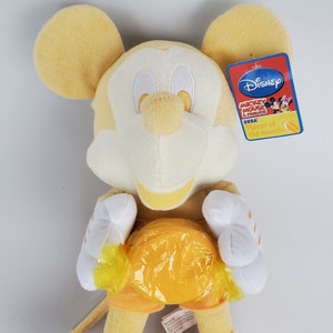 Puede incluir: Juguete de peluche amarillo de Mickey Mouse sosteniendo un caramelo amarillo. El juguete lleva un atuendo amarillo y blanco y tiene una cara blanca con ojos negros. El juguete tiene una etiqueta Disney Mickey Mouse and Friends Flavor of the Month adjunta.