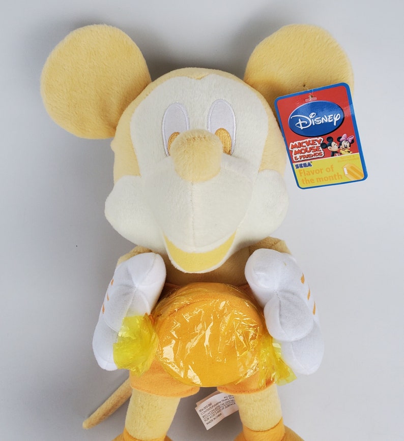 Puede incluir: Juguete de peluche amarillo de Mickey Mouse sosteniendo un caramelo amarillo. El juguete lleva un atuendo amarillo y blanco y tiene una cara blanca con ojos negros. El juguete tiene una etiqueta Disney Mickey Mouse and Friends Flavor of the Month adjunta.