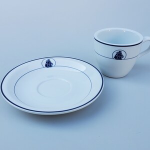 May include: A white porcelain teacup and saucer with a blue trim and a ship design. The ship design is on both the cup and the saucer.