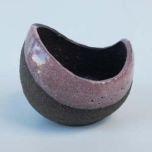 May include: A small, ceramic bowl with a dark brown exterior and a pink interior. The bowl is shaped like a crescent moon and has a textured surface.