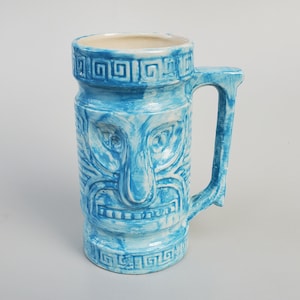 May include: A blue ceramic mug with a tiki face design and a white glaze. The mug has a handle and a geometric pattern around the top and bottom.