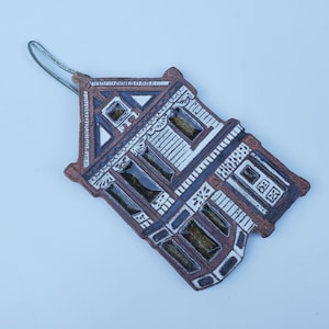 May include: A ceramic ornament shaped like a house with brown and white glaze. The house has a detailed facade with windows and a door.