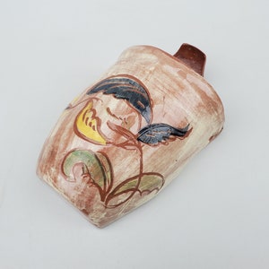 May include: A brown ceramic vase with a floral design in black, yellow, and green. The vase has a unique shape and a slightly textured surface.