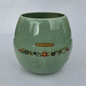 May include: A pale green ceramic cracker jar with a floral band and the word "CRACKERS" on a label. The jar is round with a textured finish. The floral band features red, blue, and yellow flowers.