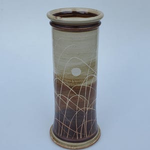 May include: A tall, cylindrical ceramic vase with a wide rim and base. The vase features a gradient of colors, transitioning from light beige to dark brown. White, curved lines and a white circle decorate the surface, creating a natural, artistic design.
