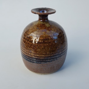 May include: A small, round, brown ceramic vase with a wide, flat opening. The vase has a brown and blue glaze with a speckled texture.