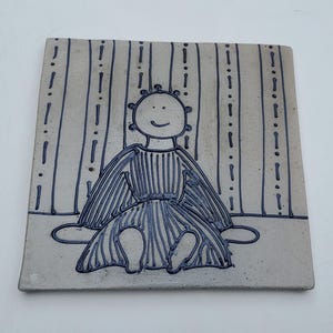 May include: A ceramic tile with a hand-drawn illustration of a person with a smiling face and a striped dress. The person is sitting in front of a striped background.