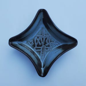 May include: A dark blue ceramic dish with a diamond shape. The dish has a design of a drum with the Roman numeral XV and a sword.
