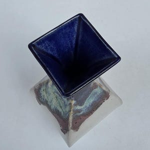 May include: A ceramic vase with a square base and a square opening. The top portion is a deep blue, while the base has a mix of colors including white, green, and brown. The vase is likely handmade and has a unique, artistic design.