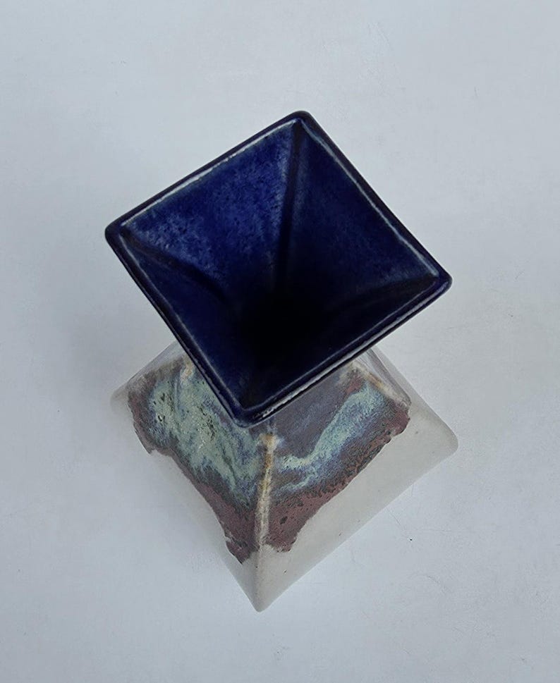 May include: A ceramic vase with a square base and a square opening. The top portion is a deep blue, while the base has a mix of colors including white, green, and brown. The vase is likely handmade and has a unique, artistic design.