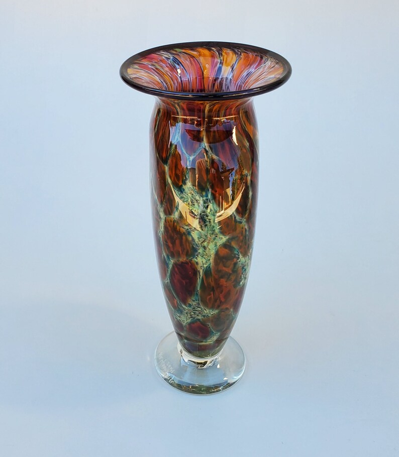 May include: A tall, brown glass vase with a wide mouth and a clear glass base. The vase has a mottled pattern of brown, orange, and green.