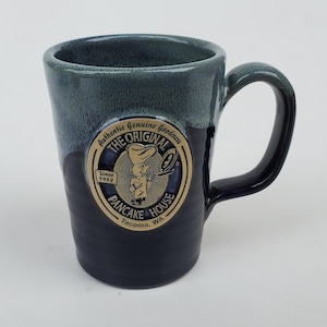 May include: A ceramic mug with a dark blue exterior and a lighter blue rim. The mug features a circular emblem with the text "The Original Pancake House" and a cartoon chef. The mug has a curved handle.