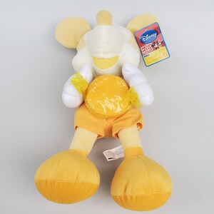 Puede incluir: Juguete de peluche amarillo de Mickey Mouse sosteniendo un caramelo amarillo. El juguete lleva pantalones cortos naranjas y tiene una cara blanca con una sonrisa amarilla. La etiqueta dice "Disney Mickey Mouse Clubhouse &iexcl;Divi&eacute;rtete mucho!"