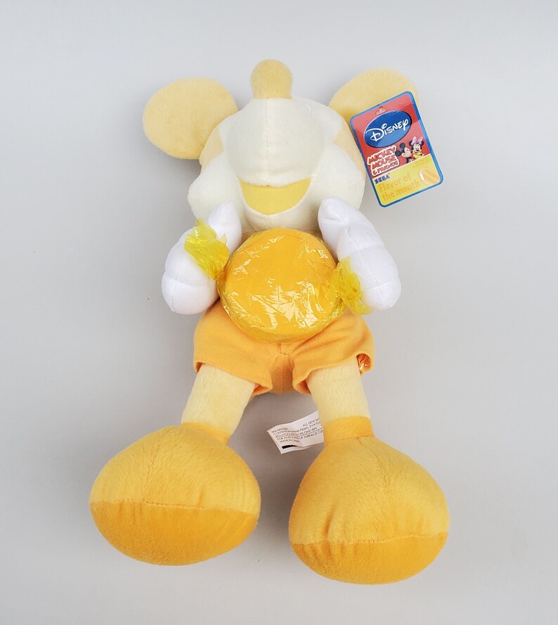 Puede incluir: Juguete de peluche amarillo de Mickey Mouse sosteniendo un caramelo amarillo. El juguete lleva pantalones cortos naranjas y tiene una cara blanca con una sonrisa amarilla. La etiqueta dice "Disney Mickey Mouse Clubhouse &iexcl;Divi&eacute;rtete mucho!"
