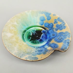 May include: A ceramic plate with a unique, abstract design. The plate features a swirling pattern of blue, green, and yellow colors, with a raised rim and a slightly uneven edge.