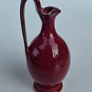 May include: A deep burgundy ceramic pitcher with a glossy finish, featuring a rounded body, a narrow neck, a flared spout, and a tall, curved handle. The edges of the handle and the rim of the spout show a subtle, textured, light-colored trim.