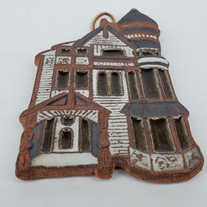 May include: A ceramic wall hanging in the shape of a house with brown, white, and black details. The house has a tower and multiple windows.