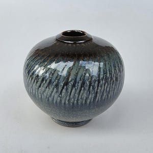 May include: A round ceramic vase with a narrow opening, featuring a glossy dark brown glaze at the top that transitions into a textured, mottled blue and grey finish with vertical streaking patterns across the spherical body of the vessel.