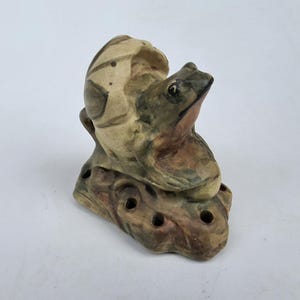 May include: A ceramic frog figurine with a mottled green and brown finish, perched atop a textured base featuring several small circular openings. The frog is positioned in front of a rounded, shell-like structure with carved line details.