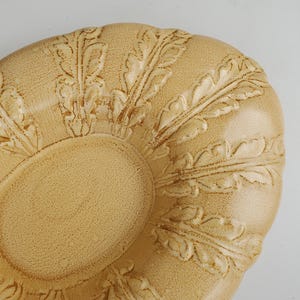May include: A beige, decorative ceramic plate with an oval center and a scalloped edge. The plate features a raised leaf pattern around the edge and center. The plate has a textured surface and a matte finish.