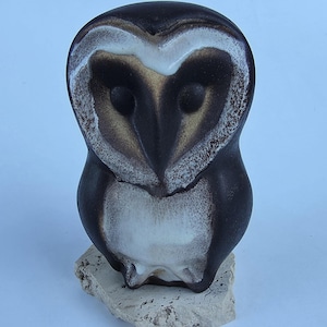May include: A ceramic owl figurine featuring a dark brown and black glaze with a white heart-shaped facial disc and chest area. The owl has a smooth, rounded body and is mounted on a rough, light-colored, porous stone base with an irregular shape.