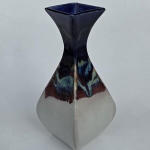 May include: A ceramic vase with a square top and a diamond-shaped body. The top is dark blue, transitioning to a mix of red, blue, and green, and then to a white base. The vase has a modern, geometric design.